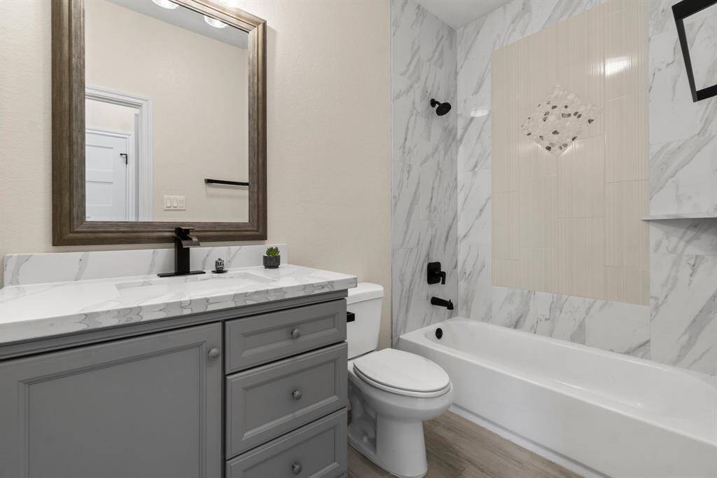 This bathroom features a modern design with a marble-style tub surround and a sleek vanity with a stone countertop. It includes a large framed mirror and contemporary fixtures, offering a clean and elegant look.