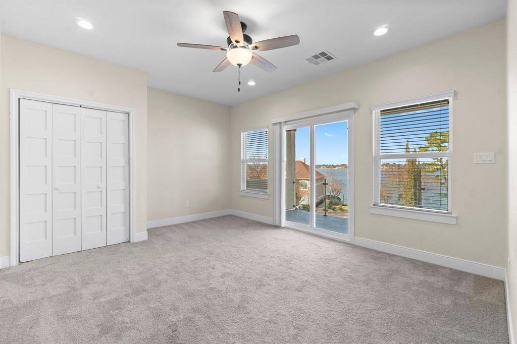 This spacious, carpeted room features a ceiling fan, ample natural light from large windows, and sliding glass doors opening to a scenic water view. It also includes a double-door closet for storage.