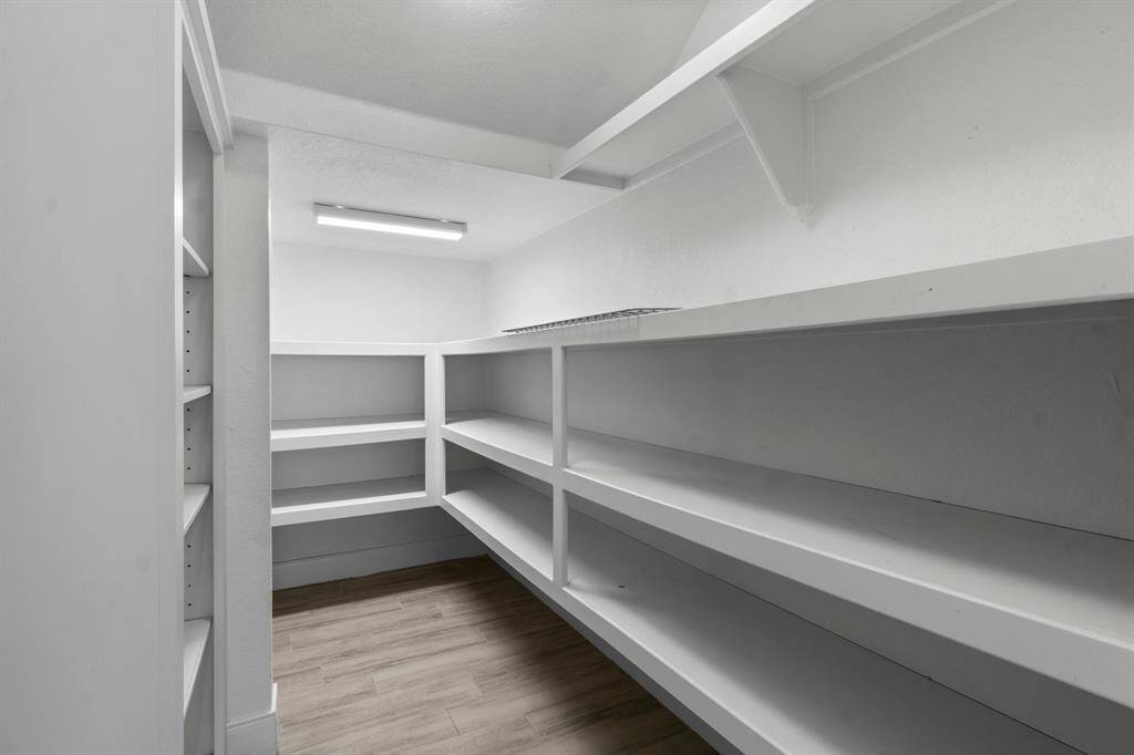 This photo showcases a spacious walk-in pantry featuring ample shelving on both sides for organized storage. The room has a clean, white finish and modern lighting, complemented by wood-style flooring. Perfect for keeping your kitchen essentials neatly arranged.