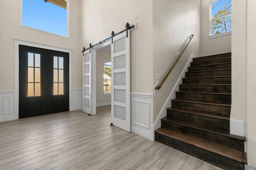 This photo showcases a stylish entryway with double doors, light wood flooring, and a staircase with dark wood steps. The space features a modern barn-style sliding door and high ceilings, offering a bright, welcoming atmosphere.