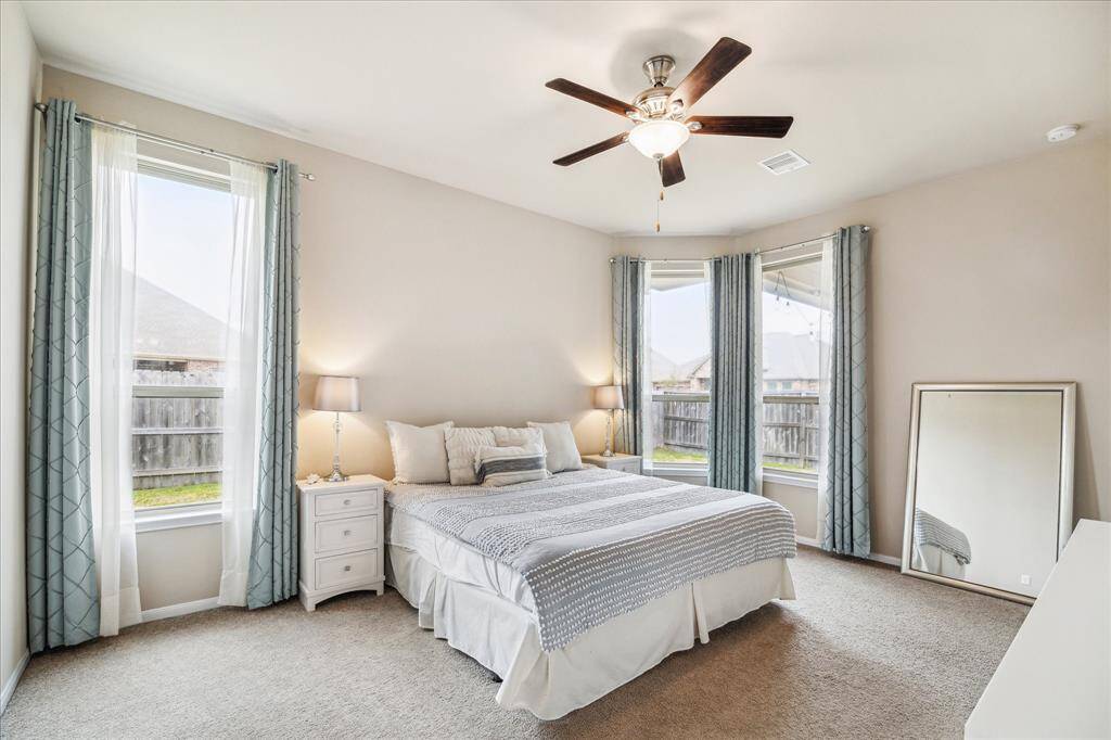 Bright and airy Primary bedroom with neutral tones, featuring large windows, plush carpeting, and a ceiling fan. The room is well-lit with natural light, located at the back of the home.