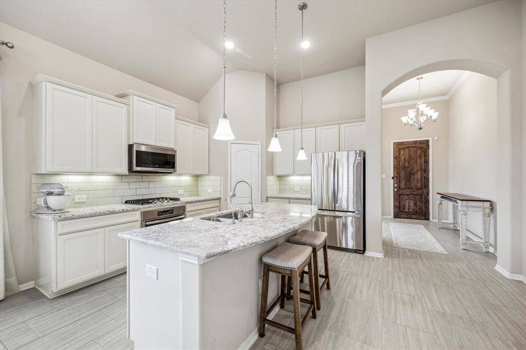 The bright kitchen features sleek white cabinetry, upgraded granite countertops, a gas range, a pantry and stainless steel appliances. It includes an island with a sink and seating space, pendant lighting, and an open layout.