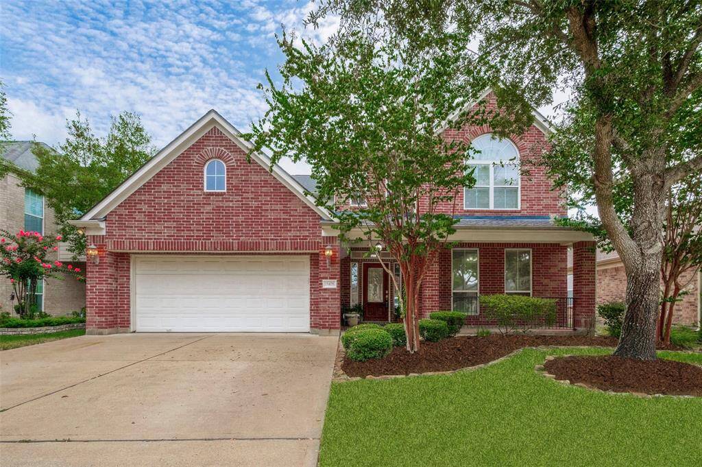 This is a two-story red brick home with a prominent gable, white trim, and an arched window above the garage. It features a well-maintained lawn, mature trees, and a welcoming covered front porch. The property includes a two-car garage and is situated in a suburban neighborhood.