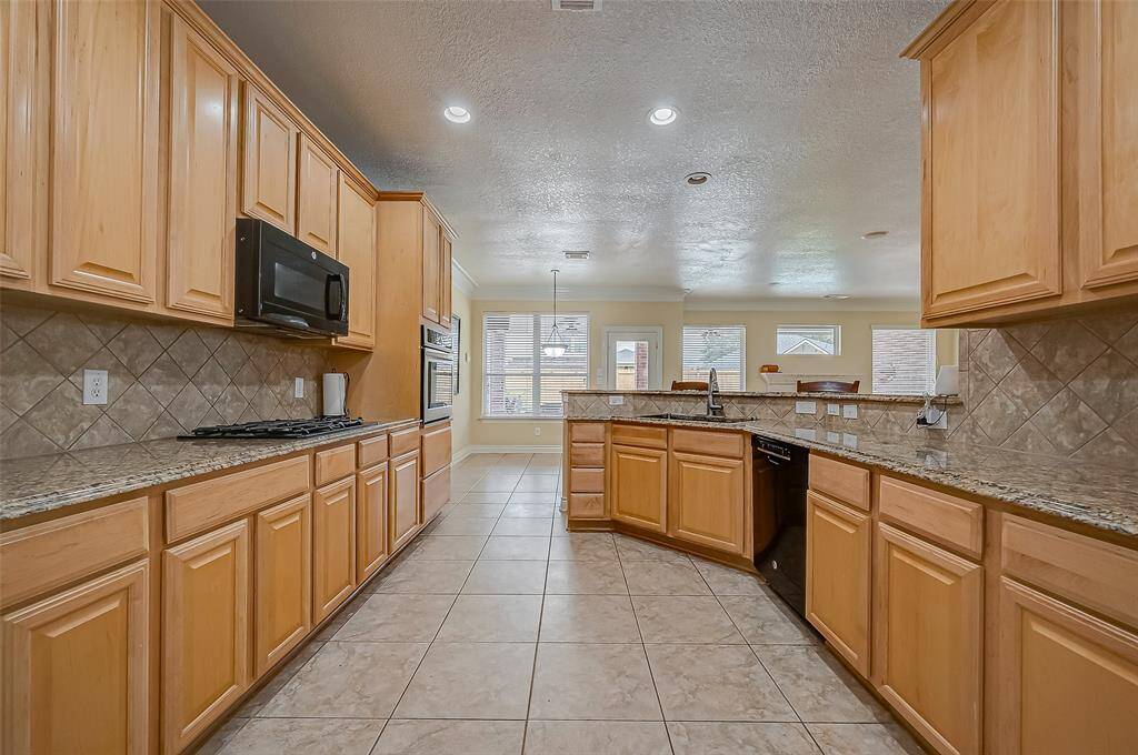 This is a spacious kitchen with ample LED overhead lighting an natural light, featuring wooden cabinetry, solid granite countertops, and modern appliances. The tiled floor and backsplash add a touch of elegance.