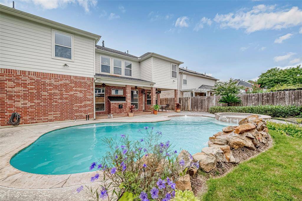 This is a spacious backyard with an inviting in-ground pool, a well-maintained lawn, and an outdoor patio area, perfect for relaxation and entertainment. The home features a brick exterior with siding and large windows.