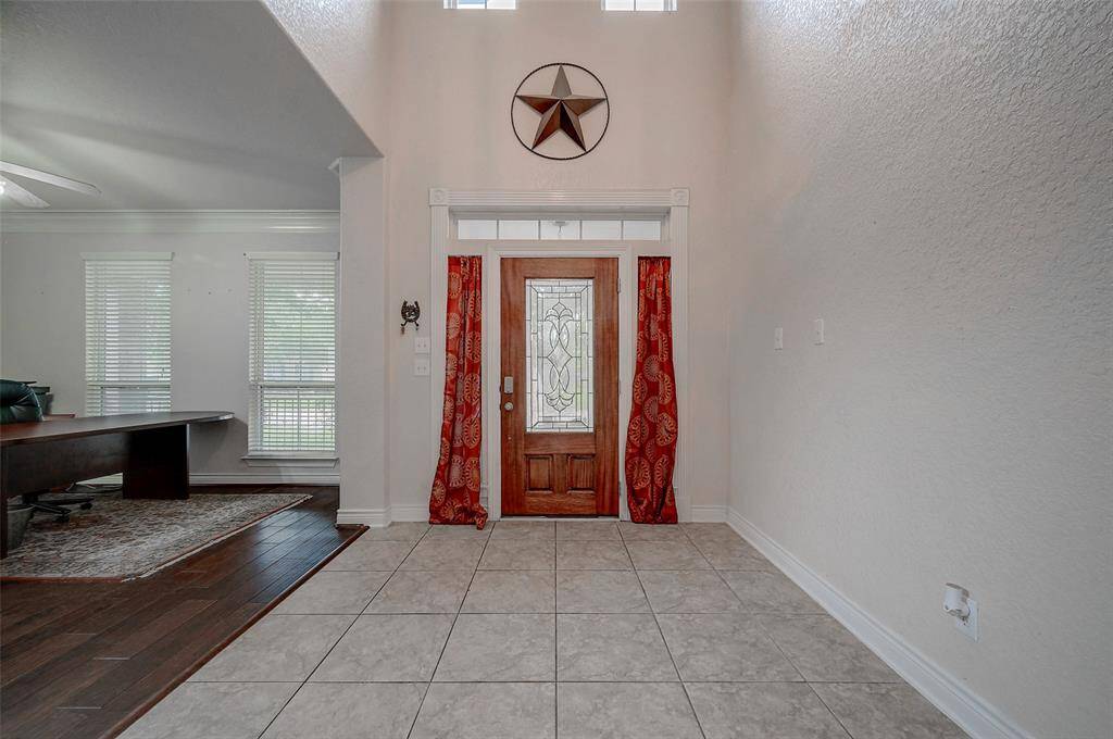 This is a spacious entryway featuring high ceilings, a decorative front door with sidelights, tile flooring, and a contrast of dark hardwood in your home office. The space is well-lit with natural light from the windows, overhead LED lighting and has a neutral color scheme.