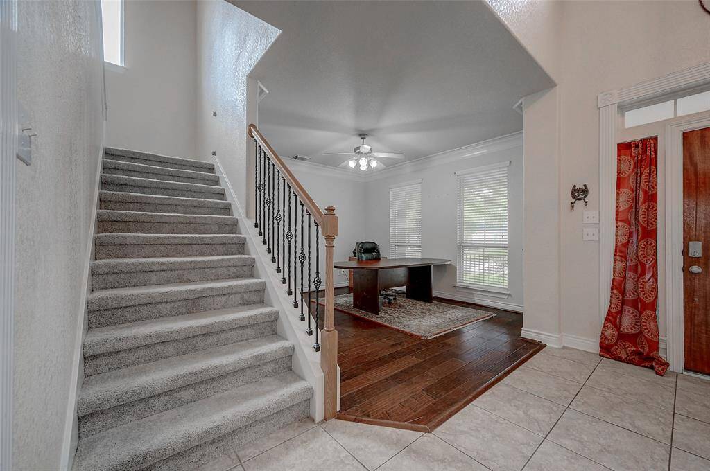 This is an inviting entryway with a carpeted staircase featuring a stylish wrought-iron railing. The space includes a combination of tiled and hardwood flooring, and is well-lit with natural light from the windows and a ceiling fan fixture. The area is complemented by neutral wall colors and has a warm, welcoming ambiance.