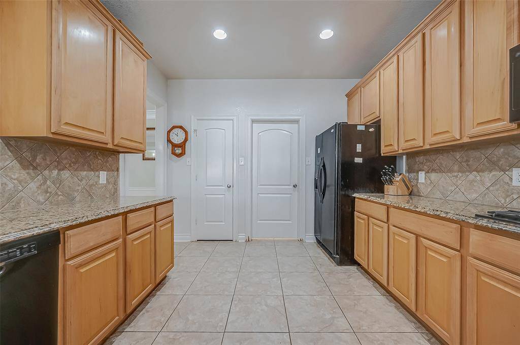 This is a modern kitchen featuring wooden cabinets, granite countertops, tile backsplash, with tile flooring. It comes equipped with black appliances, including refrigerator, and has LED recessed lighting and walk-in pantry for additional storage.