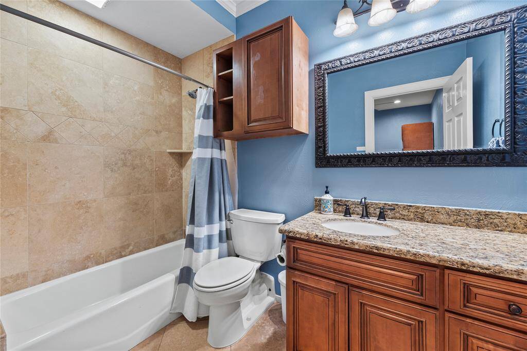 This full ensuite bathroom offers warm finishes including a furniture style granite topped vanity with a framed mirror and vanity lights above, a coordinating over-the-john cabinet, and a fully tiled tub/shower enclosure.