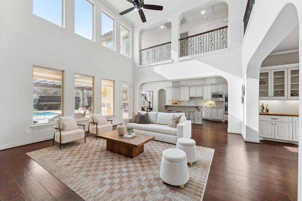 This spacious living area features high ceilings and abundant natural light from large windows. An open layout connects the living room to the kitchen, hardwood floors and a view of the pool enhance the elegance and appeal.