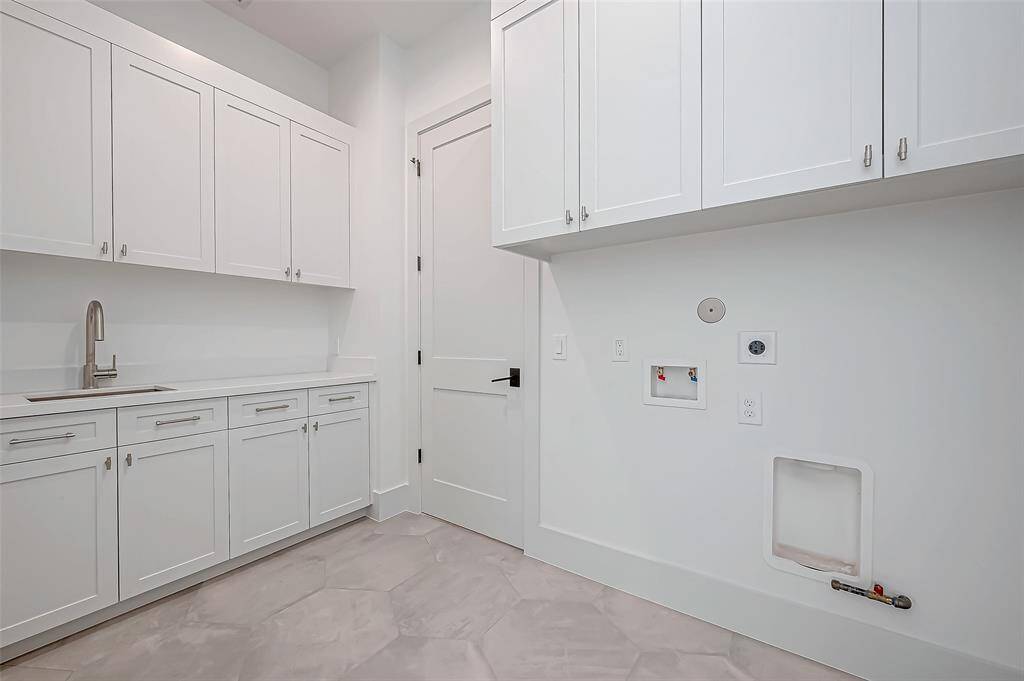 This is a bright, modern laundry room with ample white cabinetry, a sleek sink, and connections for a washer and dryer. The room features a clean, minimalist design and hexagonal tile flooring.