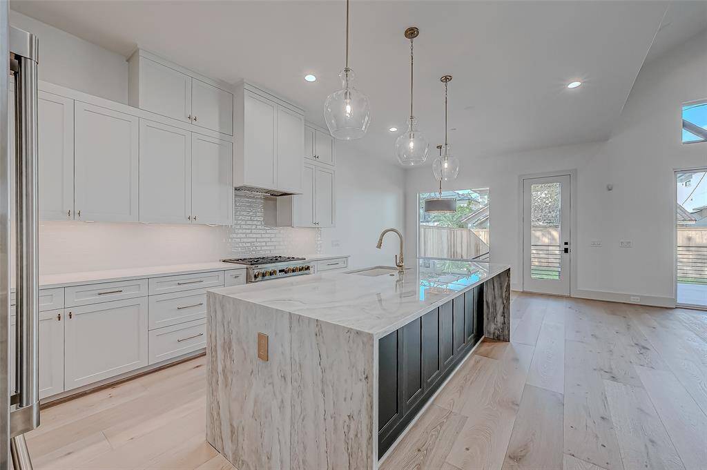 This kitchen features a sleek, modern design with white cabinetry and a large marble island. It includes stainless steel appliances, pendant lighting, and ample natural light from a nearby door and windows. The space has a clean, open layout with light wood flooring.