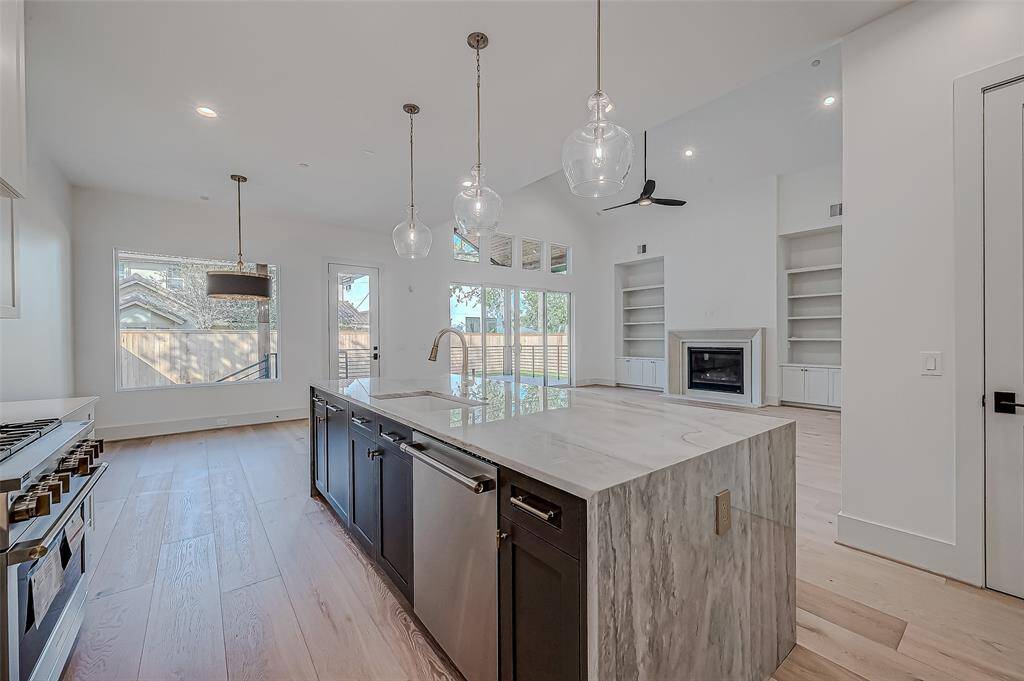 Bright and spacious open-concept kitchen and living area with light wood flooring. Features a large island with marble countertop, modern pendant lighting, and stainless steel appliances. The living room includes built-in shelves and a fireplace, with large windows and glass doors leading to the backyard.
