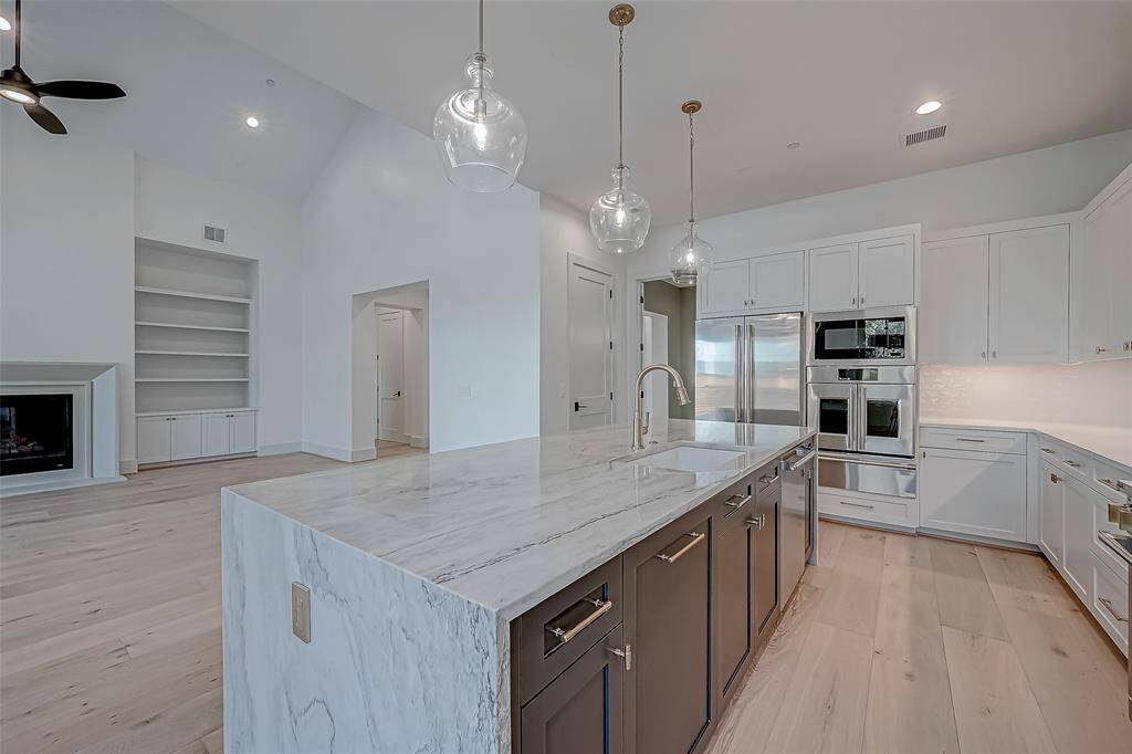 This photo showcases a modern, open-concept kitchen and living area. The kitchen features a large marble island, stainless steel appliances, and white cabinetry. The space is bright with high ceilings, pendant lighting, and light wood flooring. Adjacent is a living area with built-in shelves and a fireplace, creating a spacious and inviting atmosphere.