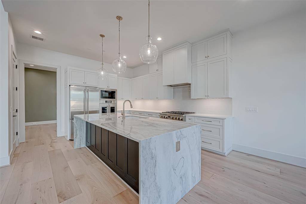 This modern kitchen features a large marble island with a built-in sink, elegant pendant lighting, and ample cabinetry. Stainless steel appliances, including a double oven and refrigerator, complement the bright, open layout. The light wood flooring adds warmth to the sleek design.