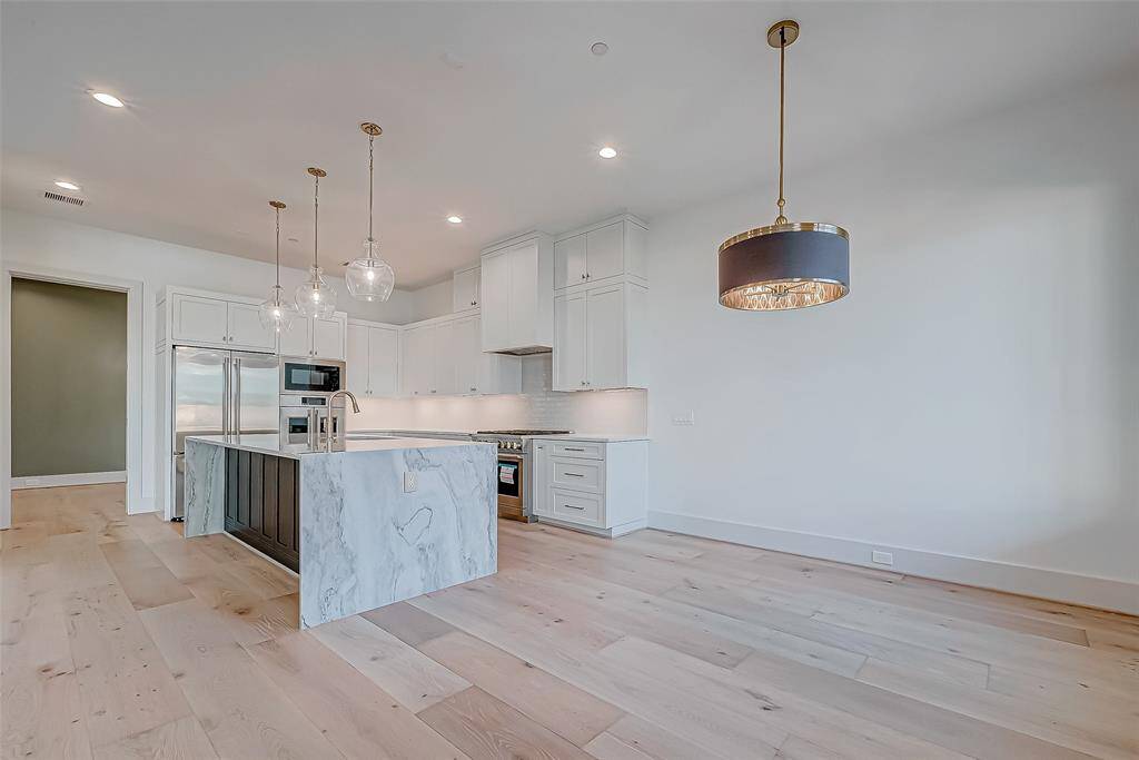 This photo showcases a modern kitchen with a spacious open layout. It features a large marble island with a built-in sink, pendant lighting, and sleek white cabinetry. Stainless steel appliances include a refrigerator, oven, and microwave. The light wood flooring adds warmth to the contemporary design.
