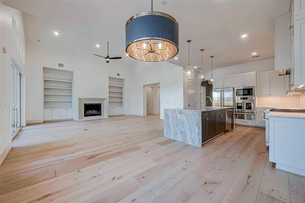 This spacious, open-concept living area features light wood flooring, a modern kitchen with a large island, and built-in shelving surrounding a fireplace. High ceilings and ample natural light enhance the airy feel, complemented by elegant lighting fixtures.