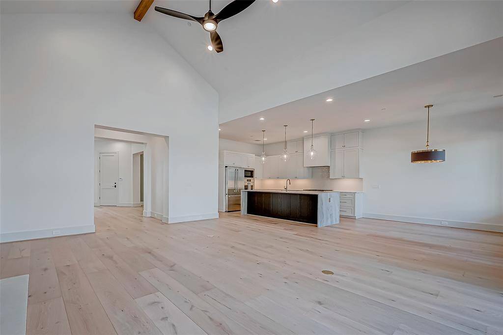 This spacious open-concept living area features high ceilings with wooden beams, modern light fixtures, and light hardwood floors. The kitchen boasts a large island with pendant lighting, sleek cabinetry, and stainless steel appliances, creating a bright and inviting space.