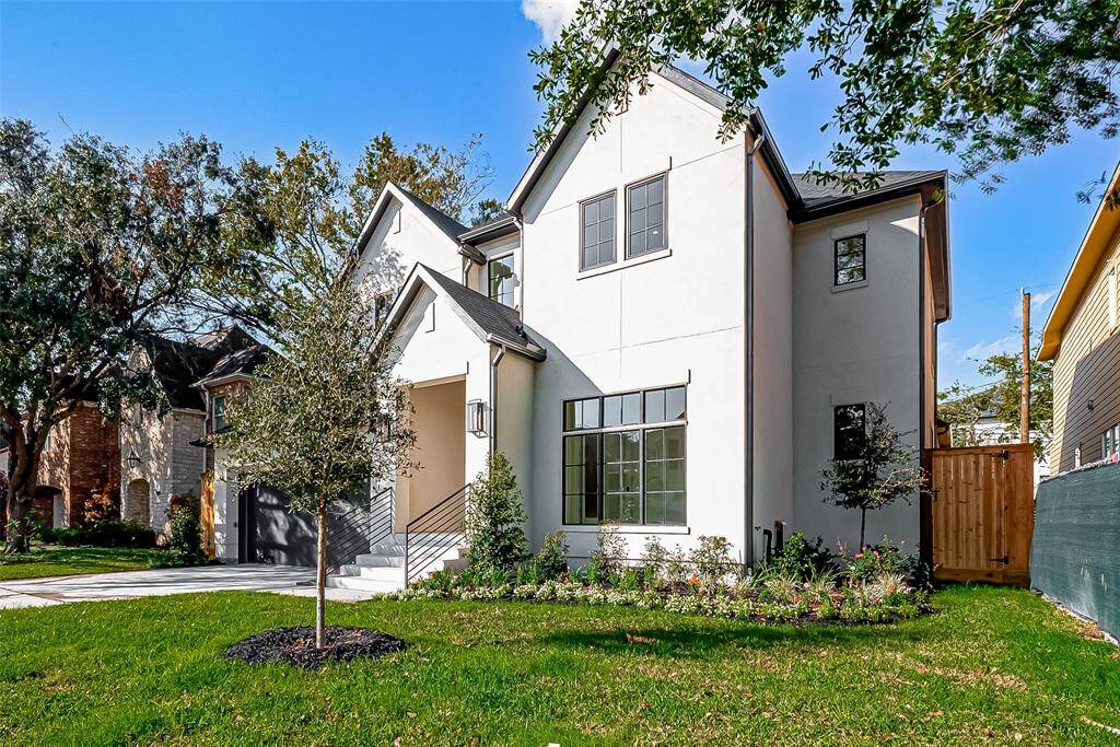 This photo showcases a modern two-story home with a clean, white exterior and dark trim. It features large windows, a well-maintained front yard with fresh landscaping, and a concrete driveway. The house has a welcoming entrance with a small covered porch and is situated in a neighborhood with mature trees.