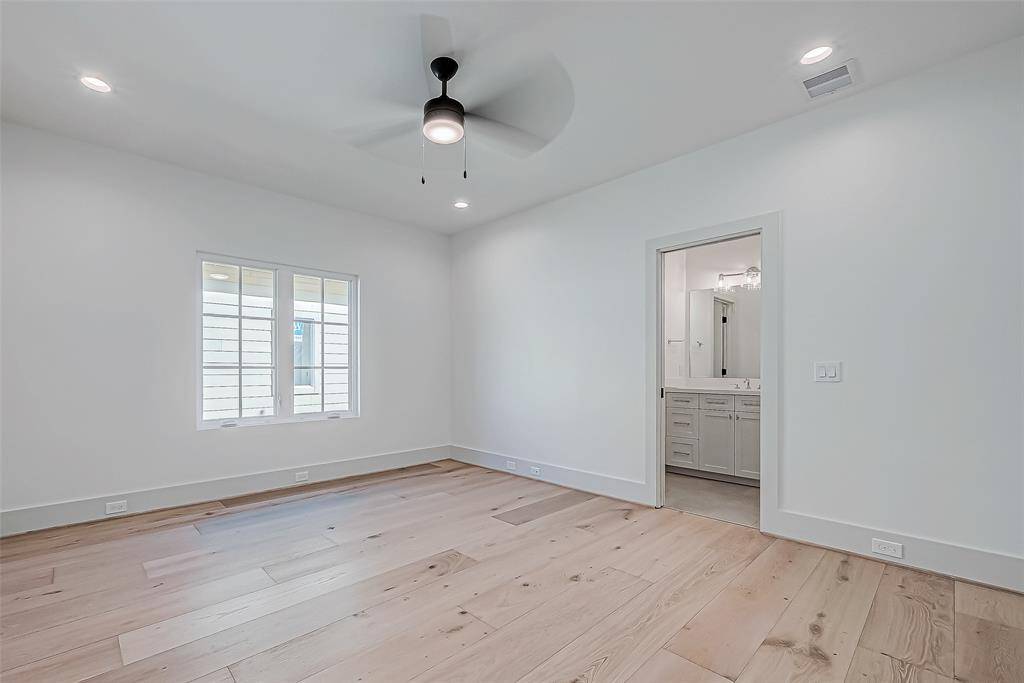 This photo features a bright, modern bedroom with light wood flooring, a ceiling fan, and recessed lighting. It includes a window with shutters and an en-suite bathroom with a sleek vanity.