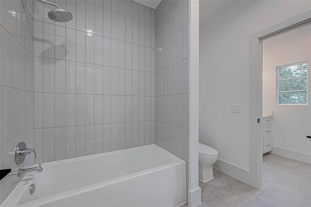 This photo shows a modern bathroom with a sleek bathtub and shower combination, featuring vertical gray tile. The space is bright with a window and includes a separate area for the toilet and a vanity with drawers. The overall look is clean and contemporary.