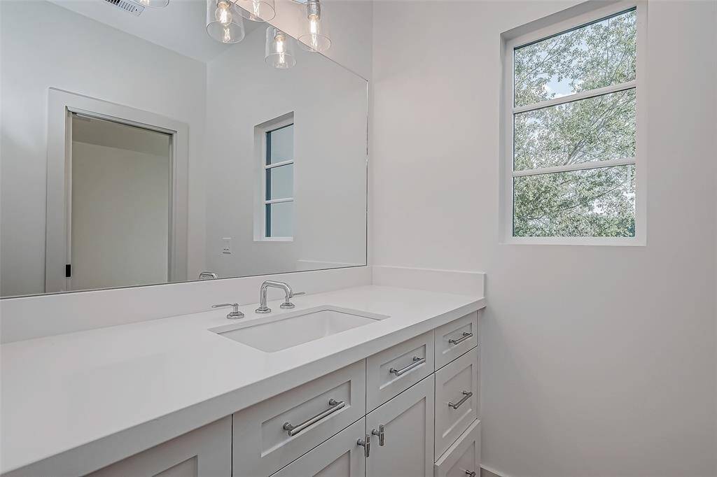 This bathroom features a modern design with a large mirror, sleek white vanity, and ample storage. Natural light streams through a window, highlighting the clean, minimalist aesthetic. A contemporary light fixture adds a stylish touch.