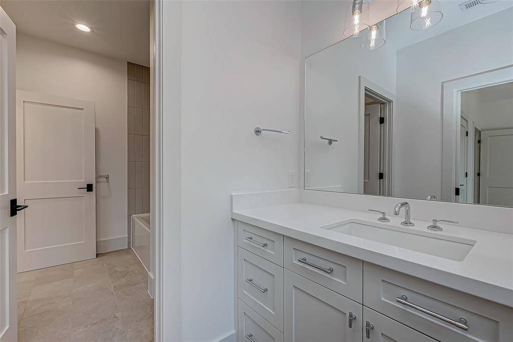 This bathroom features a modern design with a sleek white vanity, integrated sink, and ample storage. The space is well-lit with contemporary light fixtures and includes a bathtub with tiled walls.