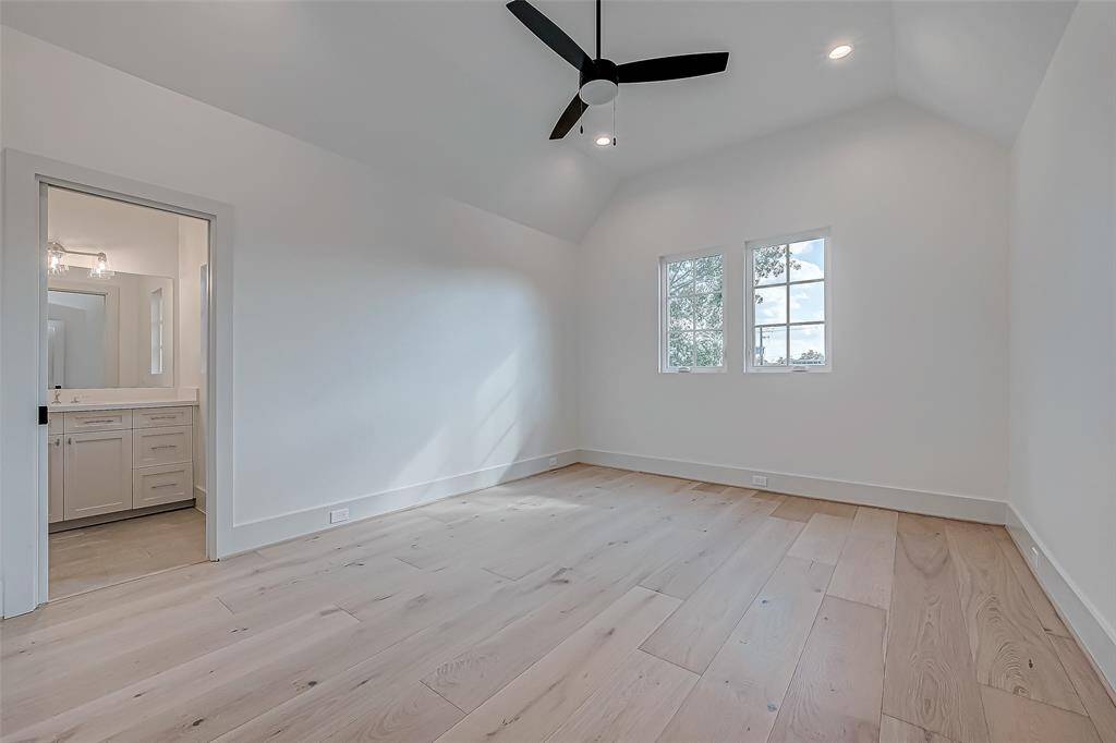 This bright, spacious bedroom features light wood flooring, a modern ceiling fan, and ample natural light from two large windows. It includes an en-suite bathroom with white cabinetry, offering a clean and contemporary feel.