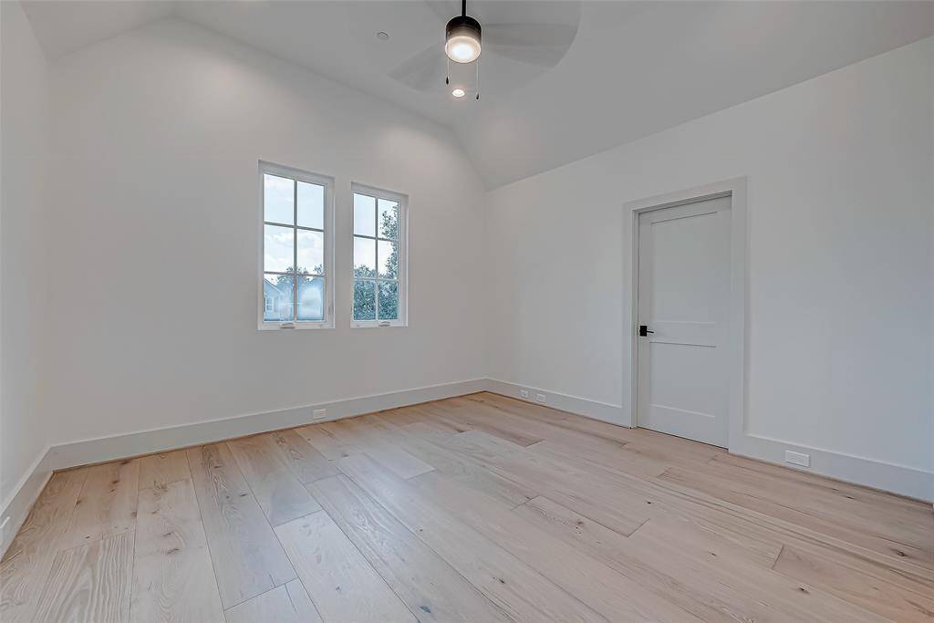 This room features high ceilings with recessed lighting and a ceiling fan. It has large windows that provide natural light, light-colored walls, and hardwood flooring. There's a single door, offering a clean and modern look.