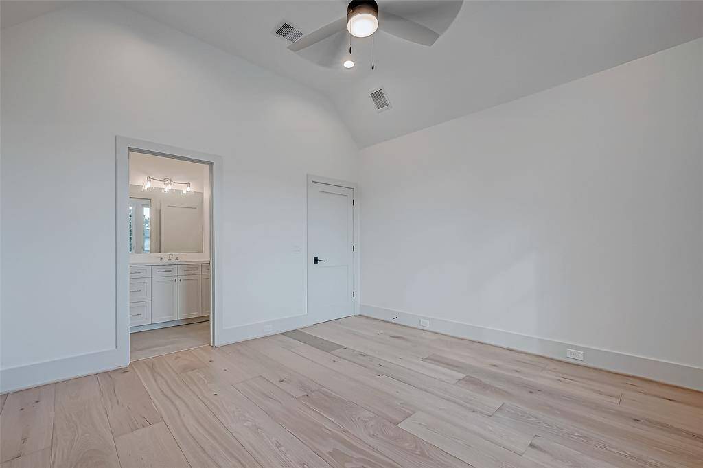 This room features light wood flooring and a ceiling fan, with an en-suite bathroom visible through an open door. The walls are painted white, creating a bright and spacious feel.