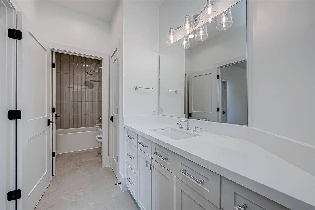 This bathroom features a modern design with a large mirror, sleek white vanity, and ample storage. The space is bright with contemporary lighting, and the adjacent room has a tiled shower and bathtub.