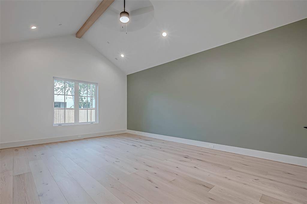This room features a vaulted ceiling with a wooden beam and a ceiling fan, complemented by light wood flooring. A large window provides natural light, and the space is accentuated by a green accent wall, creating a modern and airy atmosphere.