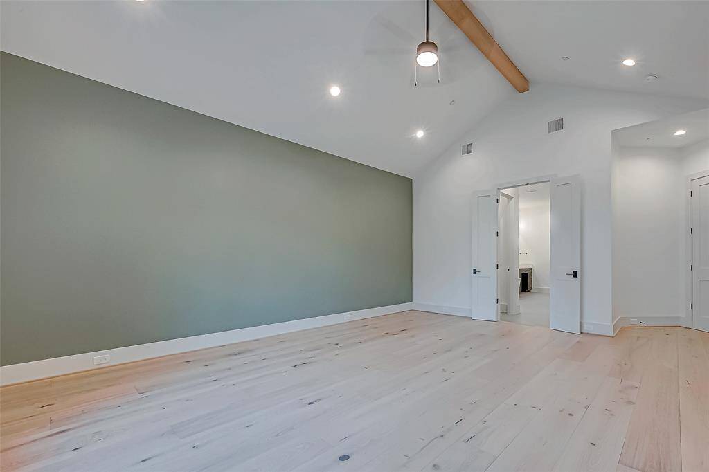 This spacious room features a vaulted ceiling with a wooden beam, recessed lighting, and a ceiling fan. It has light wood flooring and an accent wall in a soft green hue. Double doors lead to an adjoining space, adding to its open and airy feel.