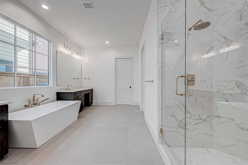 This photo showcases a spacious, modern bathroom featuring a sleek freestanding bathtub, dual vanities with ample counter space, and a luxurious glass-enclosed shower with marble-style tiles. Large windows provide natural light, enhancing the clean and elegant design.