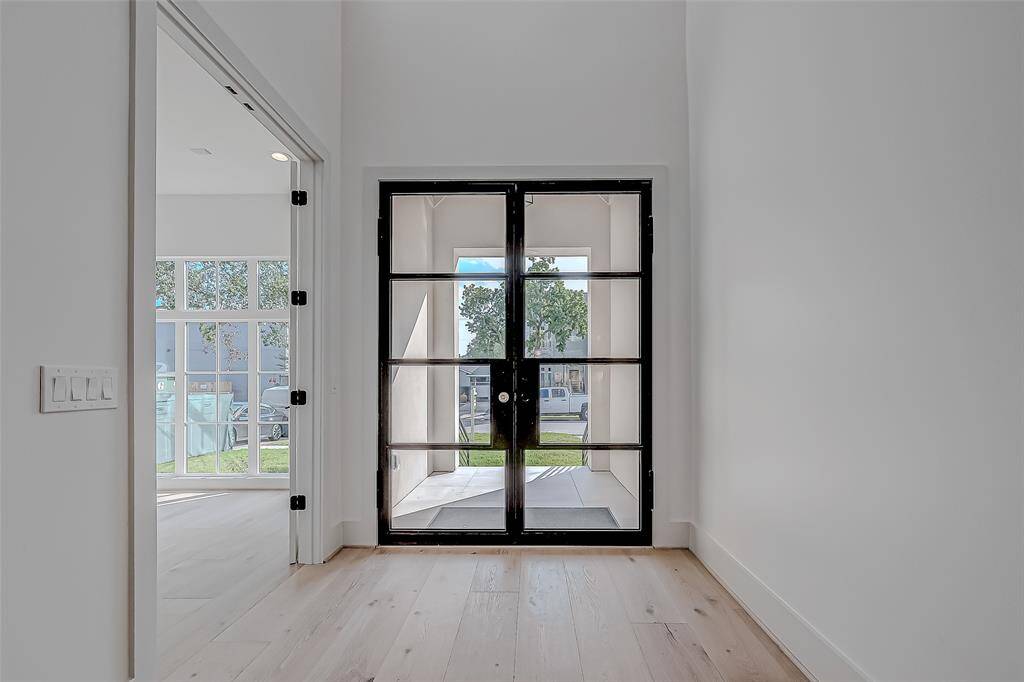 This photo showcases a bright, modern entryway with sleek black-framed glass doors opening to a view of greenery outside. The adjacent room features large windows, allowing ample natural light to fill the space. The light wood flooring adds warmth to the clean, contemporary design.