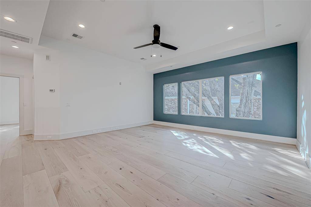 This spacious room features light wood flooring and a ceiling fan. A large window wall provides abundant natural light and a view of the outdoors, complemented by a dark accent wall. The room's neutral tones offer a versatile space for customization.