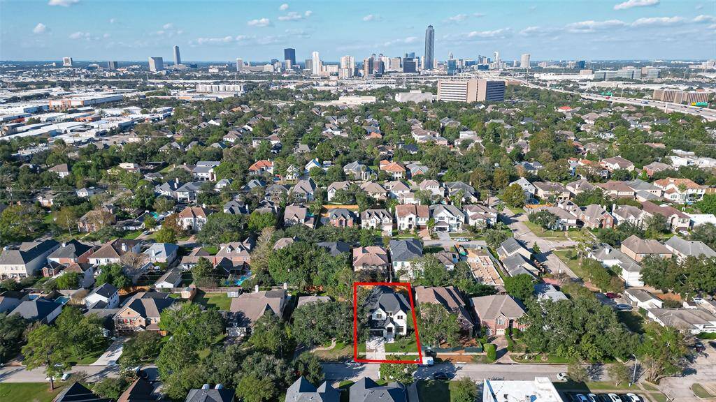 This aerial photo showcases a charming two-story home outlined in red, nestled in a lush, tree-lined neighborhood with a view of the city skyline in the distance. The area features a mix of well-maintained homes, offering a suburban feel with urban amenities nearby.