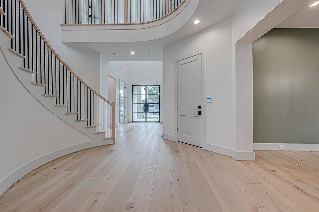 This photo showcases a modern entryway with light wood flooring, a sleek staircase with black railings, and a high ceiling. The space is bright and airy, featuring a view of a glass door leading outside.