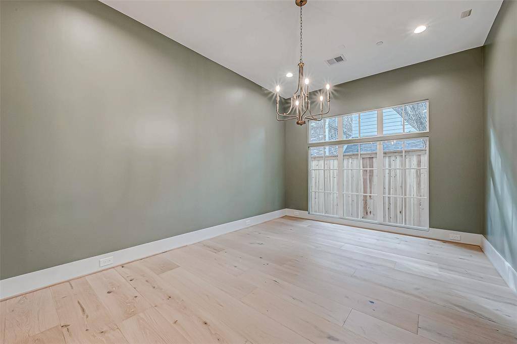 This photo features a spacious, light-filled room with light wood flooring and soft green walls. It has a modern chandelier and large windows overlooking a fenced yard, creating a bright and inviting atmosphere. Ideal for a dining or living area.