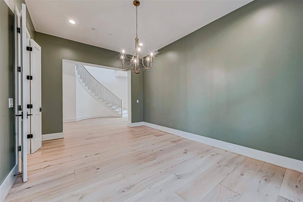 This photo features a spacious room with light wood flooring and soft green walls, accented by a modern chandelier. The room opens into a bright area with a curved staircase, adding an elegant touch. Ideal for a dining or living space with a contemporary feel.