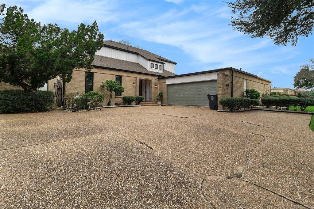 This photo showcases a modern home entrance with a welcoming front door framed by black metal gates. The exterior features light brick walls, elegant outdoor lighting, and a neatly trimmed shrub. A potted plant adds greenery, and the driveway leads to an adjacent garage.