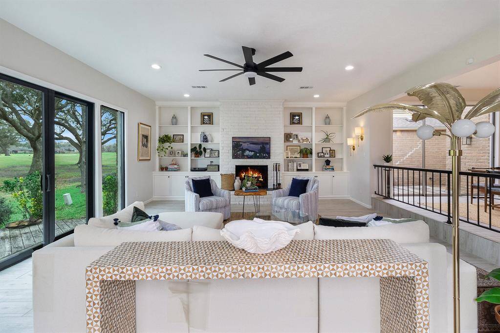 This inviting living room features a cozy fireplace, built-in shelves, and a modern ceiling fan. Large sliding glass doors offer abundant natural light and a scenic view of the lush outdoor greenery. The space is elegantly designed with a neutral color palette, perfect for relaxation and entertaining.