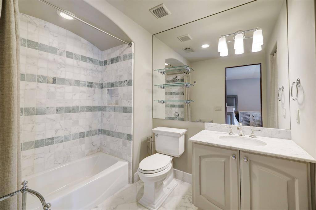 This en-suite bathroom boasts marble tile flooring and countertops, a Kohler porcelain undermount sink, a Kohler tub and shower combination with marble tile surround, a Kohler toilet, recessed lighting, and stylish mirror-mounted lampshade sconces and glass shelving.