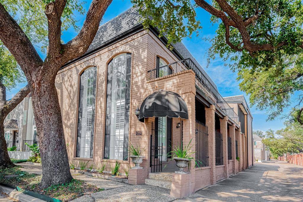 Charming corner brick home with 22' arched windows and a black awning above the entrance. The property is shaded by mature trees, adding to its curb appeal.