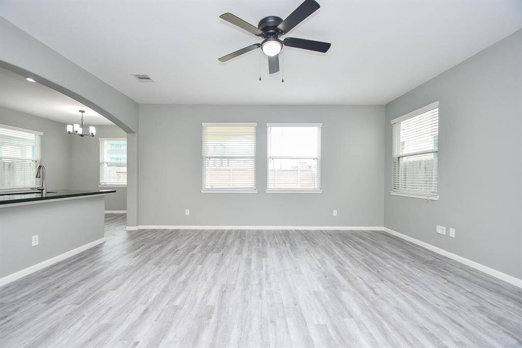 A wide, airy layout defines the living room, where soft gray walls and durable vinyl flooring create a contemporary yet comfortable atmosphere. Large windows flood the space with natural light, while the ceiling fan with its central light fixture adds both style and function.