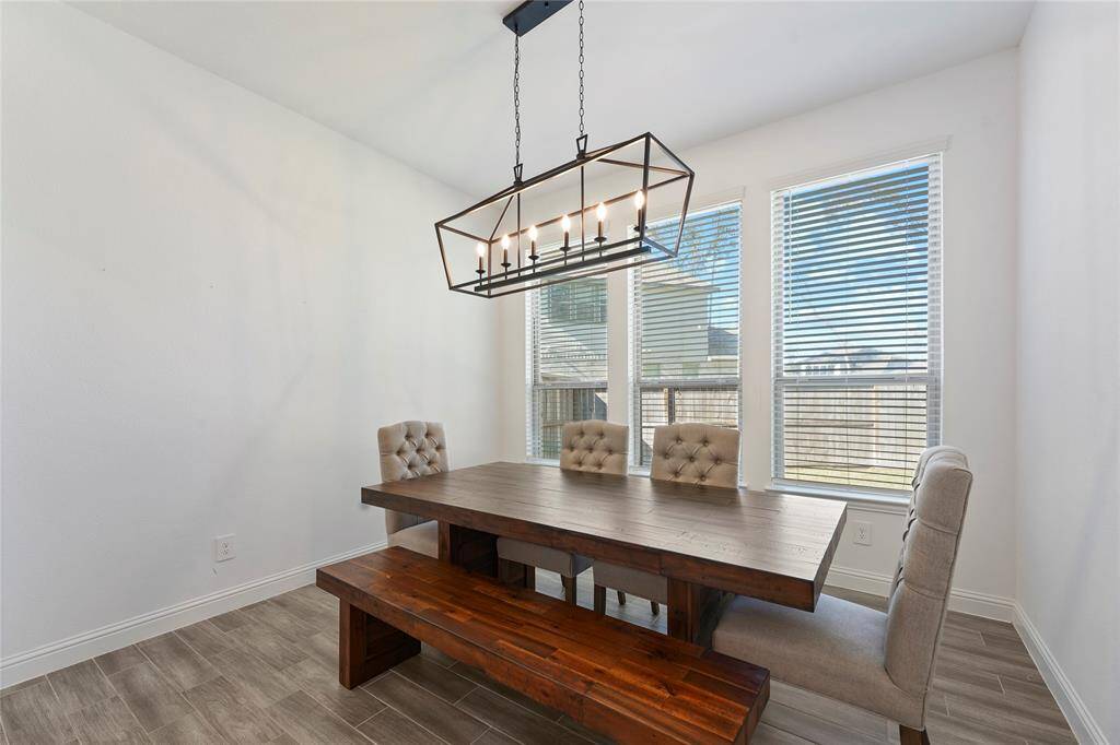 This dining area features a modern, rustic wood table with matching bench and upholstered chairs. It is well-lit by a stylish rectangular chandelier and natural light from large windows, creating a warm and inviting space.