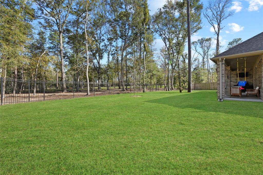 Spacious backyard with lush green lawn, bordered by tall trees and a secure fence. There's a covered patio with seating and a child's swing, perfect for outdoor relaxation and play.