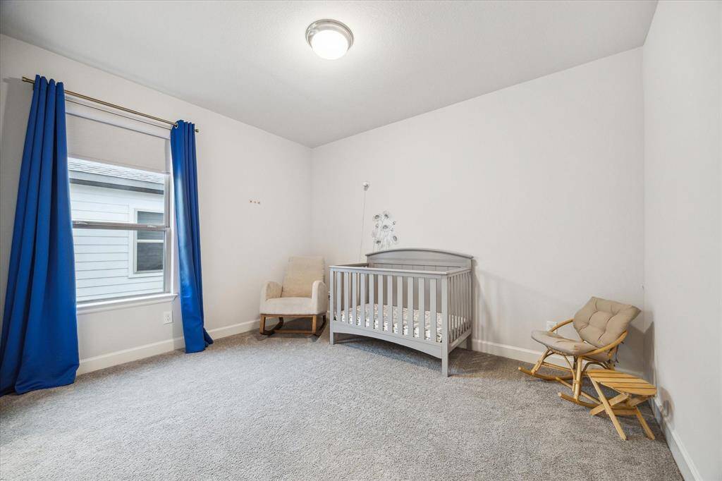 This secondary bedrooms is a cozy nursery with soft carpeting,  The room is well-lit with a window dressed in blue curtains, creating a serene atmosphere.
