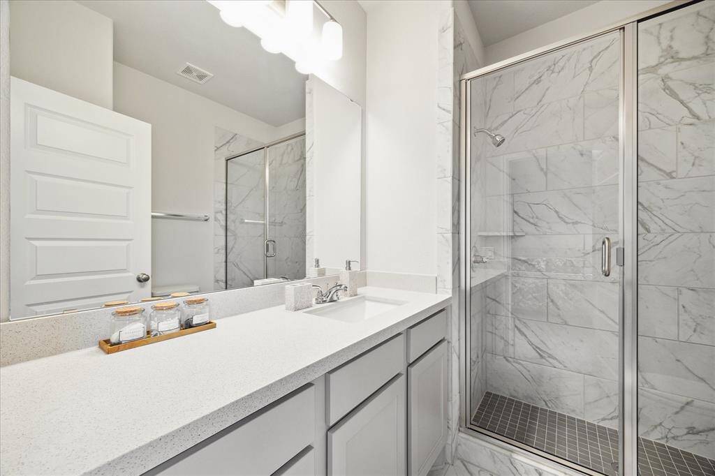 This secondary bathroom features a sleek, modern design with a spacious upgraded glass-enclosed shower showcasing elegant marble tile. It has a long countertop with dual sinks, ample storage, and contemporary fixtures, creating a bright and functional space.