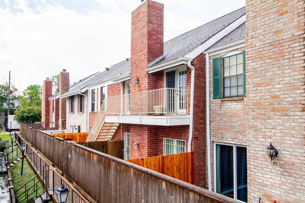 This is a multi-level, brick townhouse with a balcony and traditional design. The property includes a wooden privacy fence and exterior lighting fixtures.